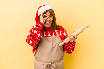 Middle age caucasian woman cooking cookies for christmas isolated on yellow background excited keeping ok gesture on eye.