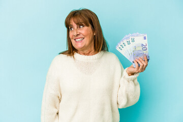 Middle age caucasian woman holding bank notes isolated on blue background looks aside smiling, cheerful and pleasant.