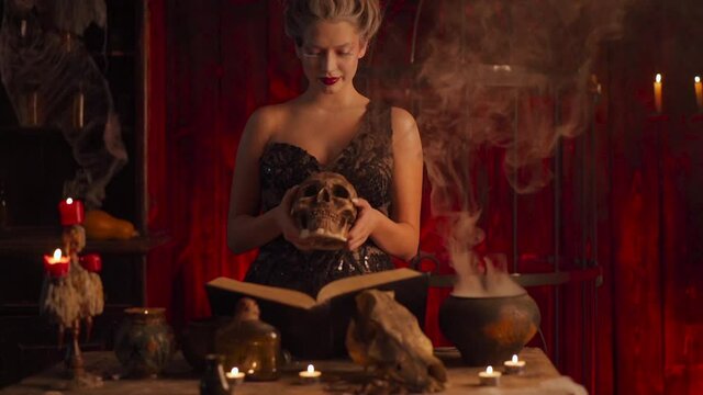 Halloween witch with cauldron and magic book. Beautiful woman flowers in hair conjuring, making witchcraft. Standing spooky dark room holding human skull in hand. Prepare love potion Handheld