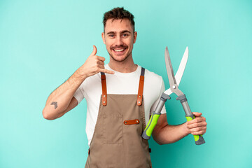 Young gardener caucasian man holding a scissors isolated on blue background showing a mobile phone call gesture with fingers.