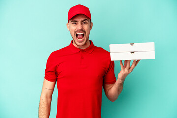 Young delivery man taking pizzas isolated on blue bakcground screaming very angry and aggressive.