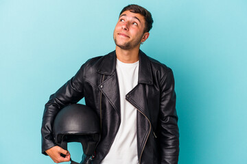 Young biker man holding helmet isolated on blue background  dreaming of achieving goals and purposes
