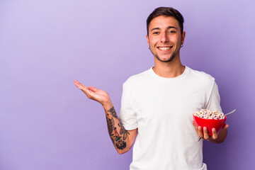 Young caucasian man eating cereals isolated on purple background  showing a copy space on a palm and holding another hand on waist.