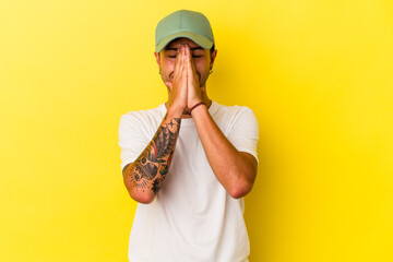 Young caucasian man with tattoos isolated on yellow background  holding hands in pray near mouth, feels confident.