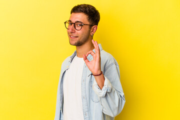 Young caucasian man with tattoos isolated on yellow background  winks an eye and holds an okay gesture with hand.