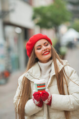 Christmas street style portrait of young beautiful fashion woman walking in European city on winter holidays, holding coffee to go. Stylish model in casual clothes with long hair is doing shopping.