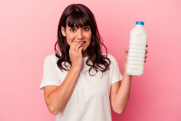 Young caucasian woman holding a bottle of milk isolated on pink background biting fingernails,...