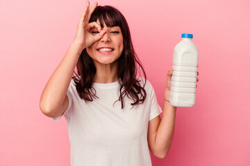 Young caucasian woman holding a bottle of milk isolated on pink background excited keeping ok gesture on eye.