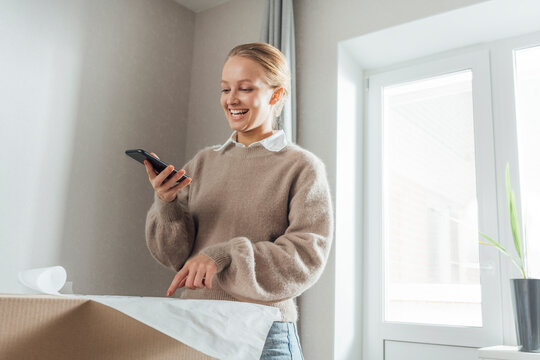 Smiling young woman with mobile phone pointing at plan on carton