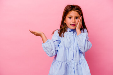 Little caucasian girl isolated on pink background  holds copy space on a palm, keep hand over cheek. Amazed and delighted.