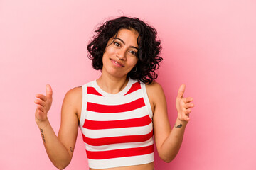 Young curly latin woman isolated on pink background holding something with both hands, product presentation.