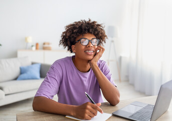 Home schooling, online education. Black teen sitting at desk with laptop, studying remotely, taking notes during class