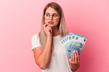 Young russian woman holding bills isolated on pink background relaxed thinking about something...