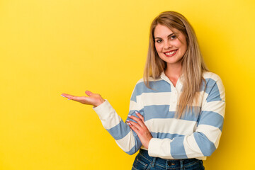 Young russian woman isolated on yellow background showing a copy space on a palm and holding another hand on waist.