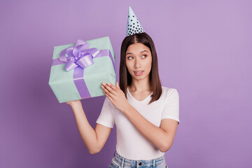 Portrait of pretty girl wearing paper cap holding in hands giftbox shake curious look empty space isolated over vivid violet color background