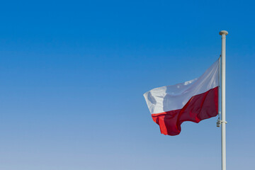 The flag of Poland flying in the wind on a tall pole against a blue sky.