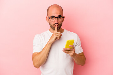 Fototapeta premium Young caucasian bald man holding mobile phone isolated on pink background keeping a secret or asking for silence.