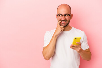 Young caucasian bald man holding mobile phone isolated on pink background  relaxed thinking about something looking at a copy space.