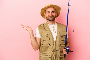 Young bald fisherman holding a rod isolated on pink background  showing a copy space on a palm and holding another hand on waist.