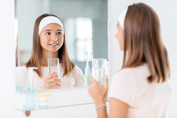 beauty, hygiene and people concept - teenage girl with glass of water looking in mirror at bathroom