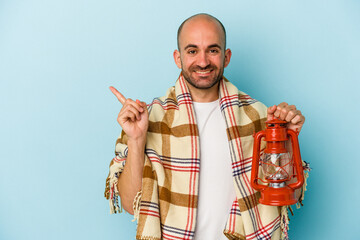 Young bald man holding vintage lantern isolated on blue background  smiling and pointing aside, showing something at blank space.