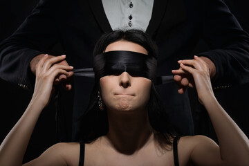 A  man closed eyes by black ribbon on the woman face and she purses lips. woman should be silent