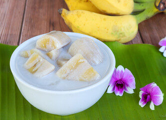 Traditional Thai dessert, bananas in coconut milk, in white bowl on banana leaf background.