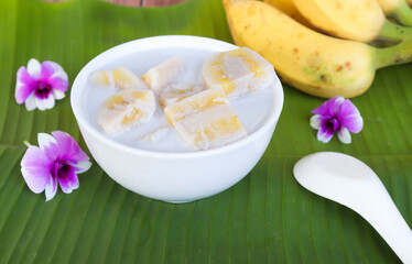 Traditional Thai dessert, bananas in coconut milk, in white bowl on banana leaf background.