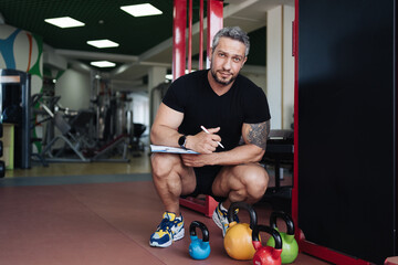 Muscular handsome trainer writing a fitness plan on clipboard for working out in the fitness