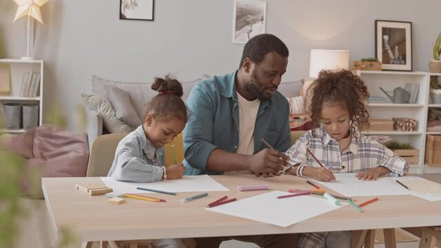 Medium slowmo shot of African-American man and his two little daughters drawing and talking spending time together at cozy home