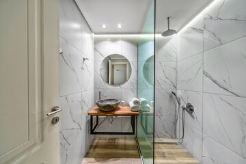 Hotel, apartment or home interior design. Standard modern bathroom with sink stone shaped on wooden stand, shower separated with glass wall, marble like tiled walles, floor laminate. 