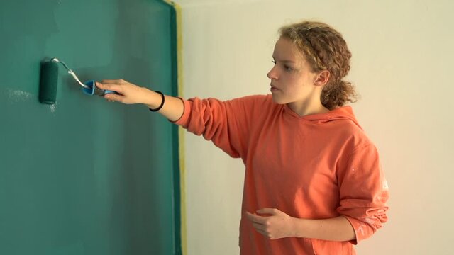 Teen girl makes a renovation in her own room. Renovation and creativity. Beautiful young girl paints the wall blue while standing on a stepladder