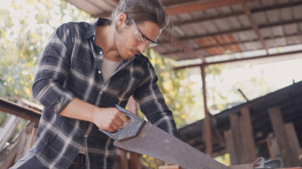 Caucasian male carpenter using hand saw for cutting wood plank at the carpentry workshop table. Craftsman making a furniture