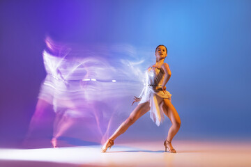 Portrait of young graceful flexible woman dancing ballroom dance without partner isolated on gradient blue purple background in neon mixed light
