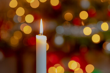 Selective focus of burning candle with blurred spotted light bokeh from Christmas tree as background, Single wax candlelight with flame and warm light in the night.