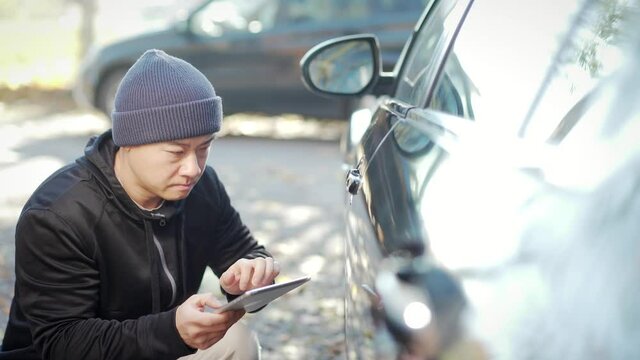 Car thief stealing a car. Hacking into the car system. Hooded tries to break the car's security with tablet. asian man standing on parking at daytime Hacking modern alarm and unlocking the door