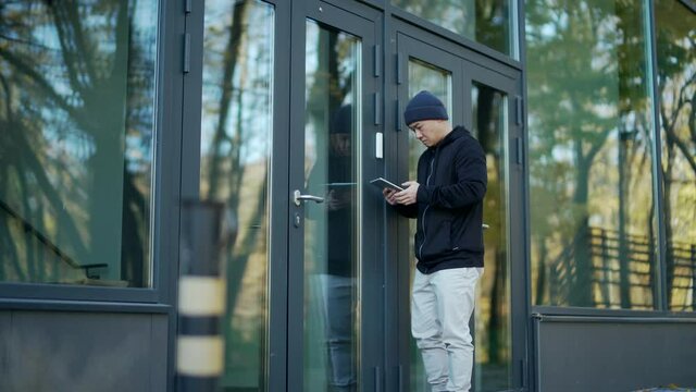 An attacker hacks into the alarm system of the entrance to a modern building or office. burglary office. thief stealing. Hooded tries to break with tablet. asian man standing Hacking, unlocking door