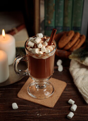 Cocoa or hot chocolate with marshmallows on a wooden table, with books, cookies, a blanket and burning candles. Vertical, low key
