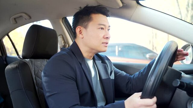 Furious angry asian man standing in a traffic jam beeps and hurries. Sitting in the car. Irritated young business male tired driver in automobile. Stressful commuting to be late work