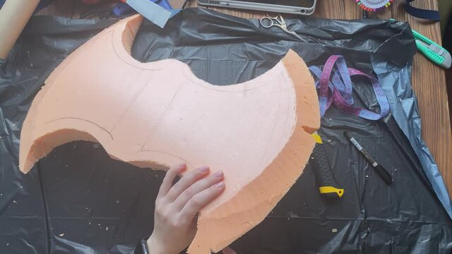 Hands cut axe form from foam polystyrene with stationery knife on garbage bag. Woman makes viking hatchet for cosplay festival. Diy celtic weapon on wooden table. Craftsman's workshop.