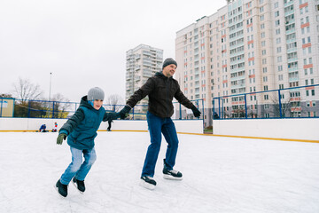 Young caucasian man hold his preschooler son arm while ice-skating at city rink in wintertime. Full lengh horizontal shot. Happy father day and winter activities concept