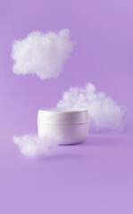 Moisturizer cream in white jar with fluffy clouds on violet background. Concept care with sensitive skin. Creative still-life photo, vertical