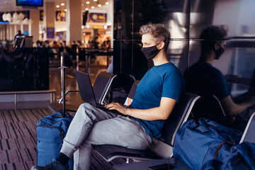 Fototapeta na wymiar Young man wearing face mask and using laptop for remote work while sitting with distance at airport. New normal, digital nomad, freelance lifestyle. Traveling during pandemic, personal protection