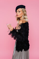 stylish woman in black beret and faux-fur jacket looking at camera while holding cocktail isolated on pink