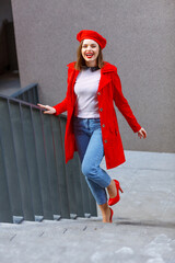 An attractive model in a red beret and a red coat walking on the stairs in a street city. Woman in jeans, brown boots. Fashion portrait, casual model goes, looks straight and smiles. Blurred details