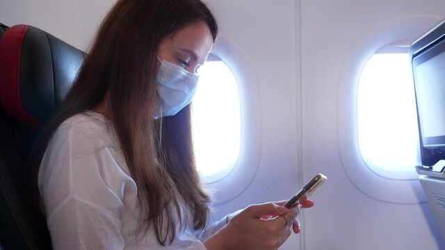Woman wearing face mask during flight, portrait shot at plane cabin. Passenger lady using smartphone, sit at economy class compartment against window