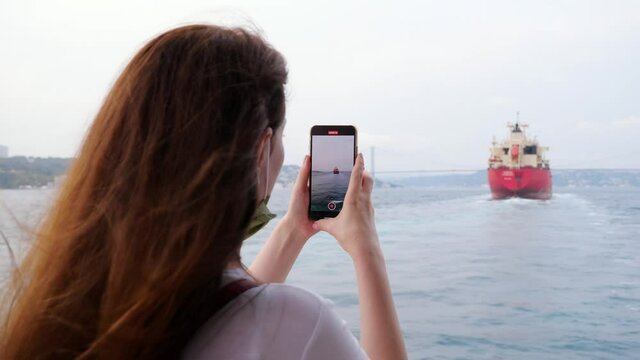 Tourist woman take photo and capture video of cargo ship sailing at Bosphorus strait. Lady uses her smartphone to capture the moments of the trip, she travels on an ordinary Istanbul ferry