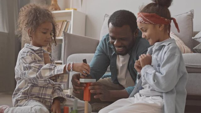 Medium slowmo shot of two cute little girls and their African-American father having fun together while playing dice game sitting on carpet next to couch in cozy living room