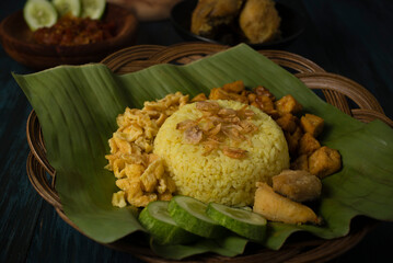 Yellow rice or turmeric rice on banana leaf and other dish like deep fried chicken, omelette, cucumber, tofu, tempe, cracker, and spicy sauces