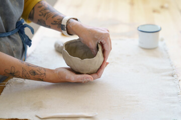 Creating handicraft pottery: closeup of working table in ceramics studio with female master shaping wet clay pot. Woman potter sculpting decorating handmade bowl in workshop, cropped shot. Art concept - obrazy, fototapety, plakaty